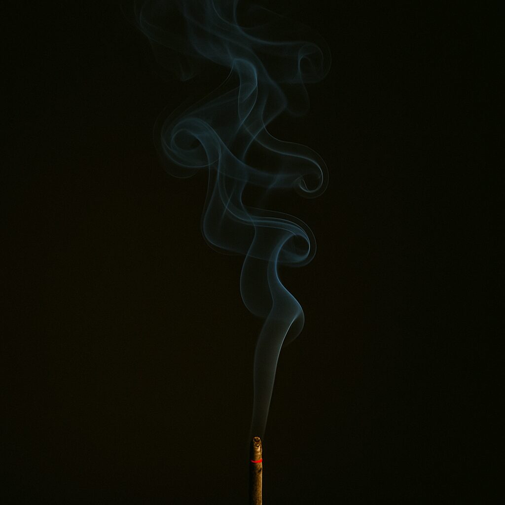 A single stick of incense burning in the dark, with thin blue smoke curling upward in soft, fluid patterns. The air feels still and meditative.